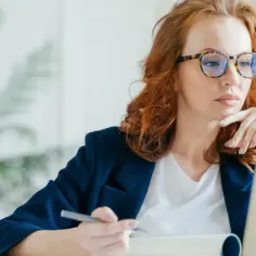 Kobieta w okularach z rudymi włosami pracuje przy laptopie, trzymając długopis.