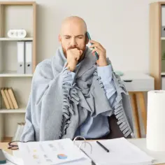 Chory mężczyzna w kocu rozmawia przez telefon, kaszląc. Widać, że jest na zwolnieniu, ale próbuje pracować.