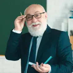 Uśmiechnięty starszy pan w okularach, z białą brodą, trzymający długopis. W tle tablica i półka z książkami. Temat: staż pracy a emerytura.