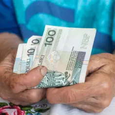 Dłonie starszej osoby trzymają banknoty 100 zł. Czy to wystarczy na życie? Najniższa emerytura w Polsce to smutna rzeczywistość.