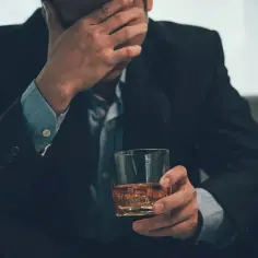 Mężczyzna w garniturze trzyma szklankę z bursztynowym płynem, zasłaniając twarz dłonią.