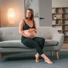 Uśmiechnięta kobieta w zaawansowanej ciąży siedzi na kanapie, delikatnie obejmując brzuch.