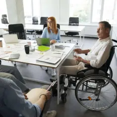 Spotkanie zespołu, gdzie mężczyzna na wózku inwalidzkim, mimo znacznego stopnia niepełnosprawności, aktywnie uczestniczy, korzystając z przywilejów dostępności.