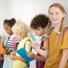 Dzieci w szkole z tabletami i telefonami, dziewczynka z warkoczami na pierwszym planie.