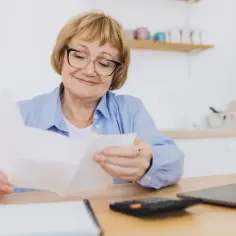Uśmiechnięta starsza pani, pracujący emeryt, analizuje dokumenty. Obok leży kalkulator i laptop, sugerując przeliczenie emerytury.