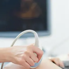Dłoń trzymająca sondę USG na brzuchu kobiety, w tle ekran monitora.