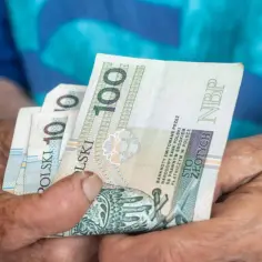 Dłonie starszej osoby trzymają banknoty 100 zł. Kto nie dostanie 14 emerytury, martwi się o przyszłość.