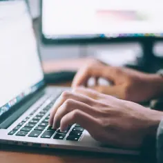 Dłonie piszące na klawiaturze laptopa, na ekranie dokument.