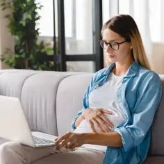 Kobieta w okularach, w ciąży, siedzi na kanapie i pracuje na laptopie. Może szukać informacji o zaświadczenie o ciąży.