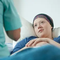 Młoda pacjentka w chustce na głowie leży w łóżku, słuchając pielęgniarki. Nadzieja na nowy lek na raka.