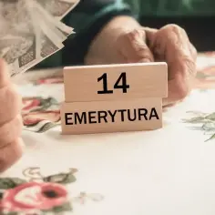 Drewniane klocki z napisem "14 EMERYTURA" na obrusie w kwiaty, obok ręki trzymającej pieniądze.